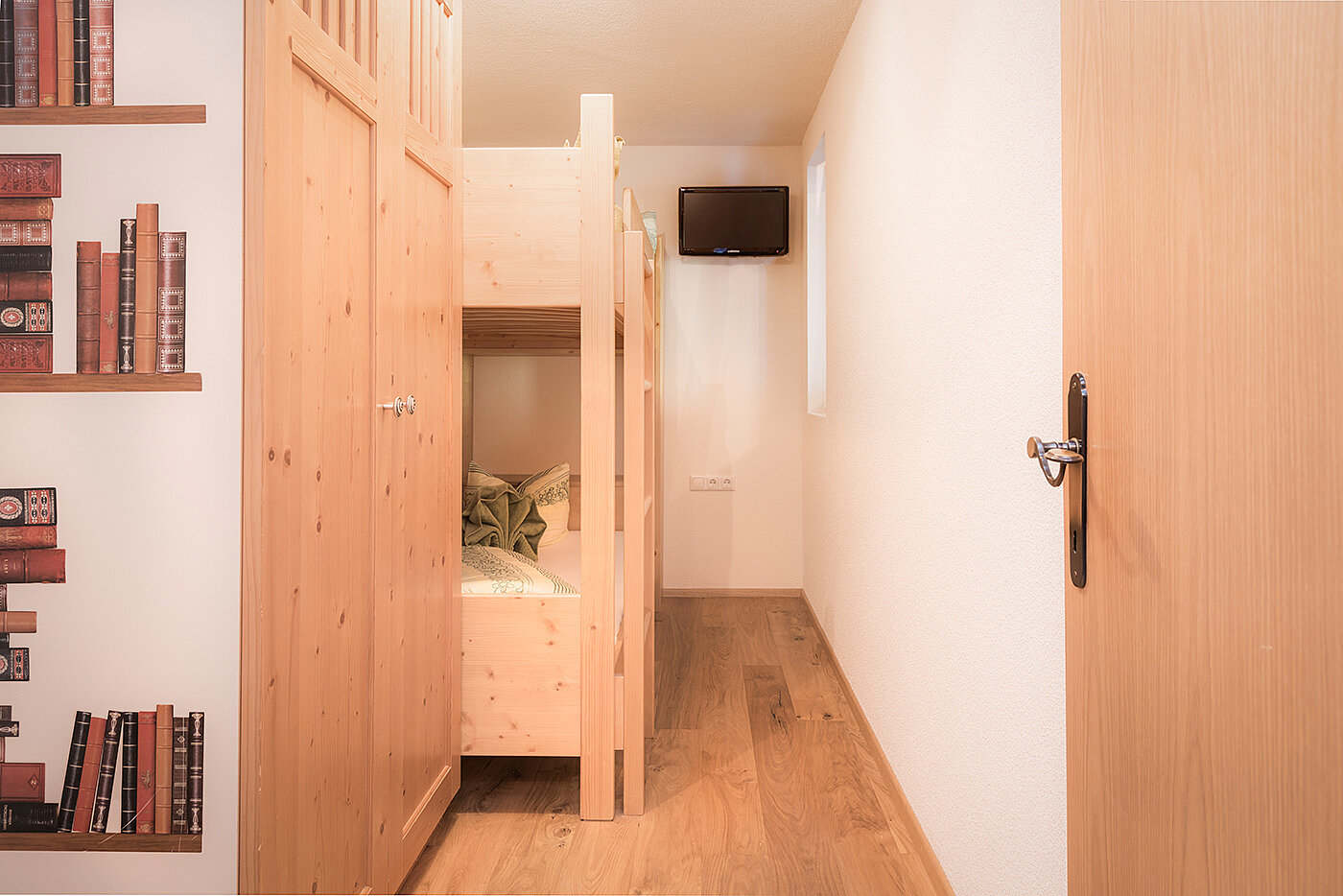 Schmales Zimmer mit Etagenbetten aus Holz, einem Bücherregal auf der linken Seite und einem kleinen an der Wand montierten Fernseher am Ende.