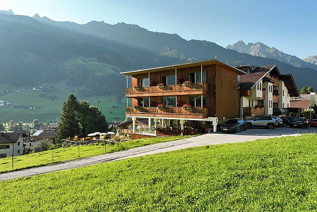 Ein modernes Holzhotel mit Balkonen liegt an einem grasbewachsenen Hang, mit Bergen und klarem Himmel im Hintergrund.