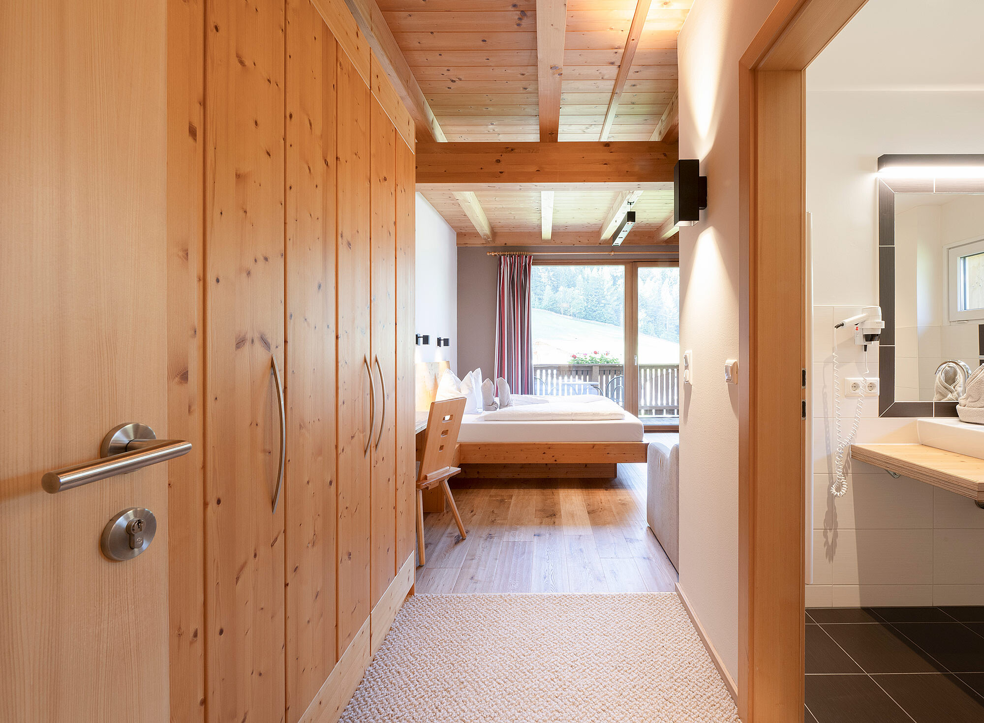 Modernes Hotelzimmer mit Holzmöbeln, Doppelbett, Schreibtisch, großem Fenster und angrenzendem Badezimmer mit beleuchtetem Spiegel.