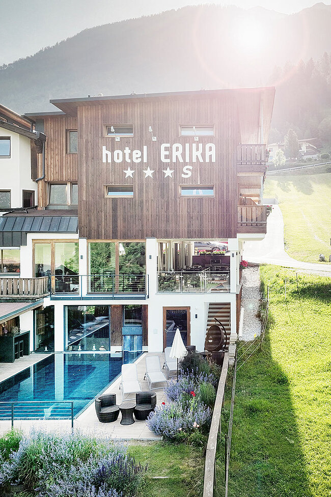 Hotel Erika mit Holzfassade, Außenpool, Sitzbereich und Bergen im Hintergrund an einem sonnigen Tag.