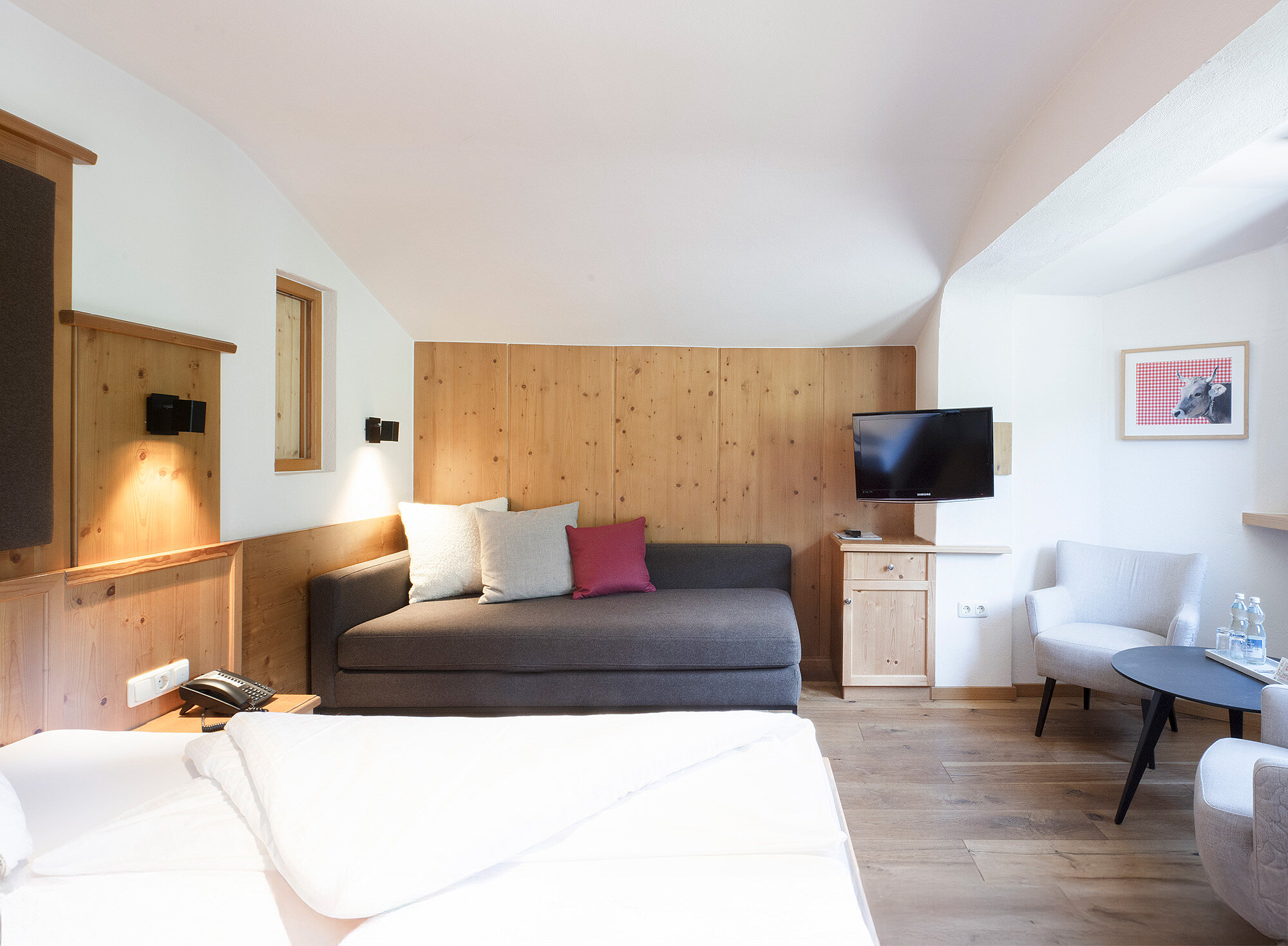 Helles Hotelzimmer mit Doppelbett, Sofa, Fernseher, Stühlen, Holzakzenten und modernem Dekor.
