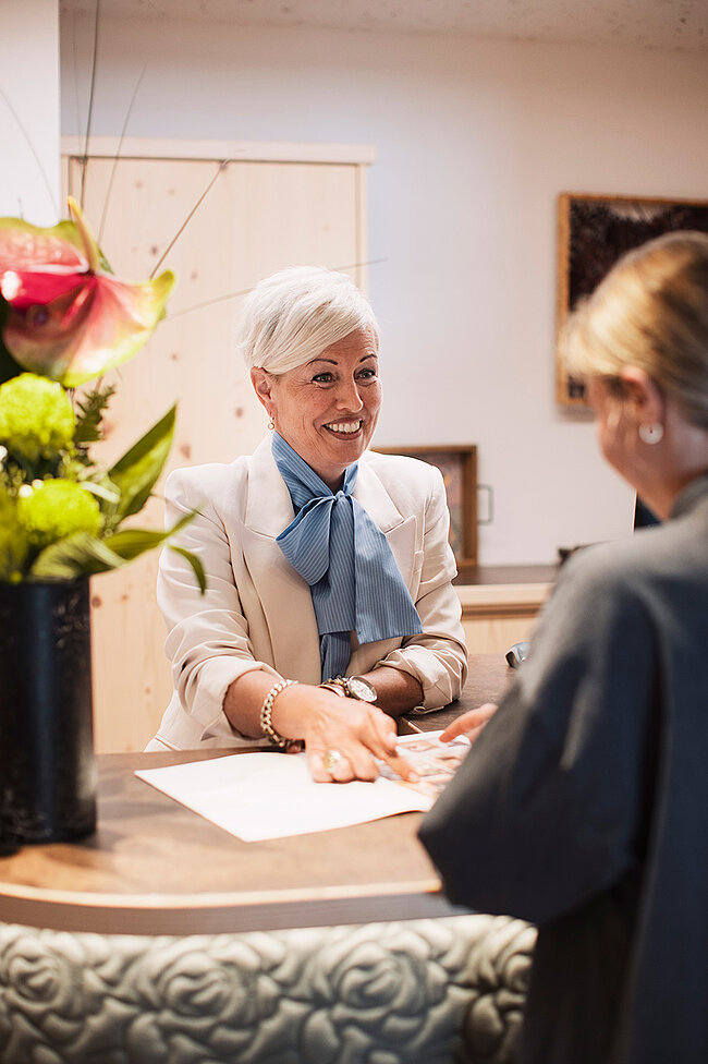 Eine lächelnde Frau mit kurzen grauen Haaren hilft einem Gast an einem Empfangstresen, in dessen Nähe Blumen stehen.