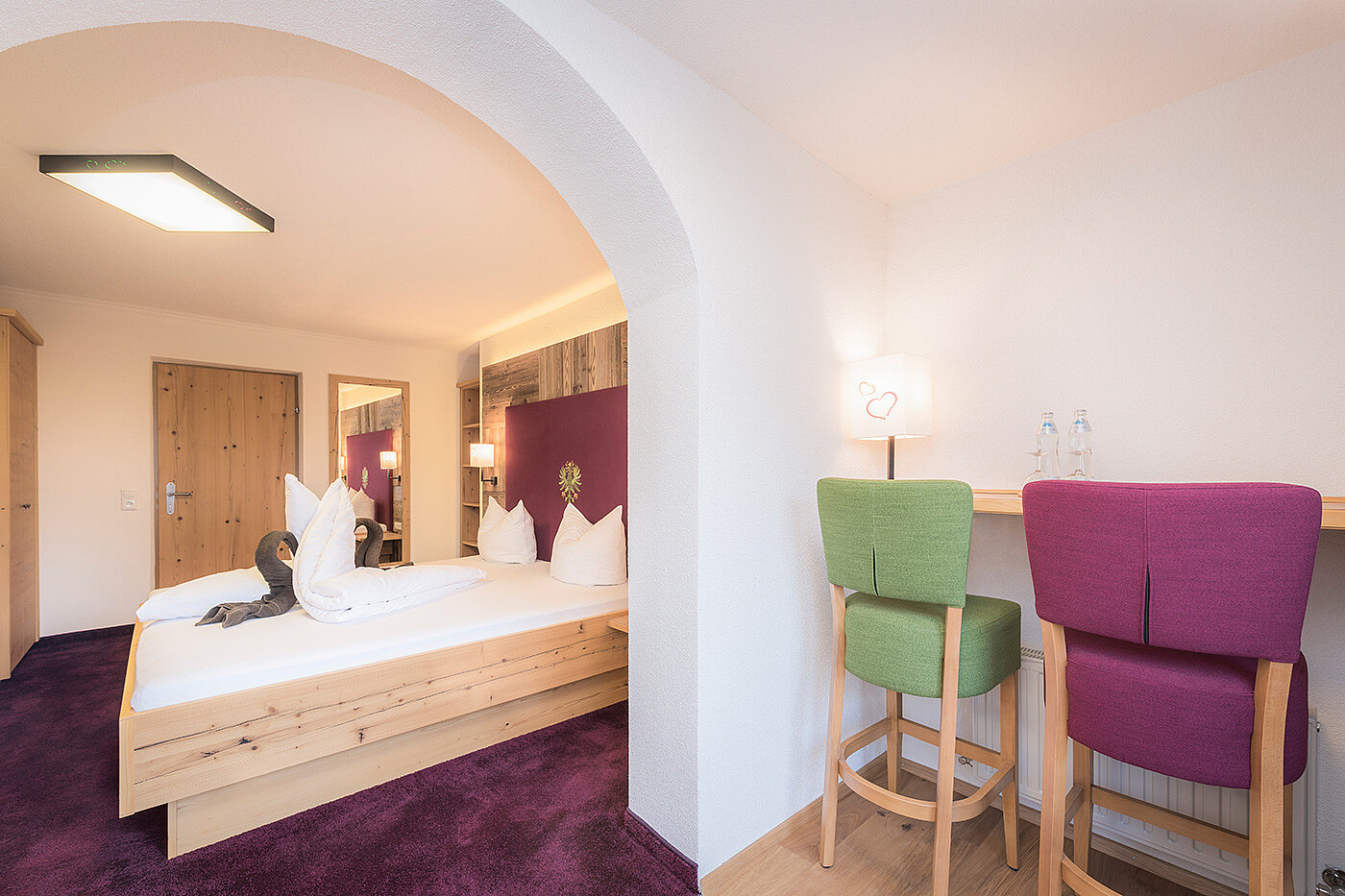  Modernes Hotelzimmer mit Doppelbett, lila Teppich und Schreibtisch mit zwei grünen und lila Stühlen.
