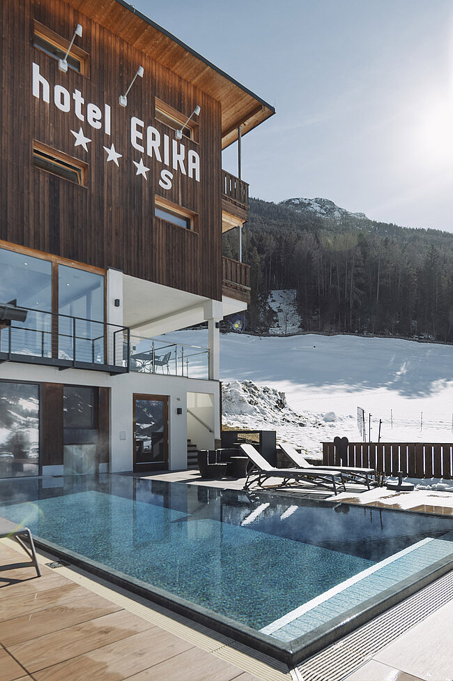 Das moderne Hotel ERIKA in Neustift überblickt einen beheizten Infinity-Pool mit aufsteigendem Dampf, Sonnenliegen und einer verschneiten Stubaitaler Berglandschaft im Hintergrund bei klarem Himmel.