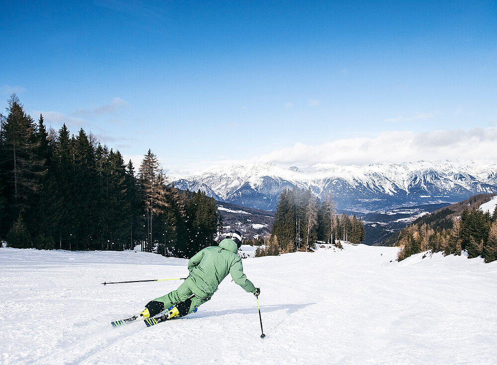 Person in grüner Skiausrüstung fährt Ski auf einem schneebedeckten Hang mit Kiefern und Bergen im Hintergrund.