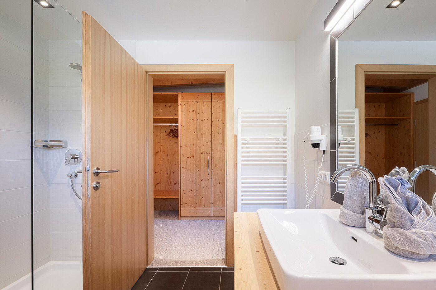 Modernes Badezimmer mit einer Holztür, die zu einem hölzernen Schrankbereich, einem weißen Waschbecken und einer sichtbaren Duschkabine aus Glas führt.
