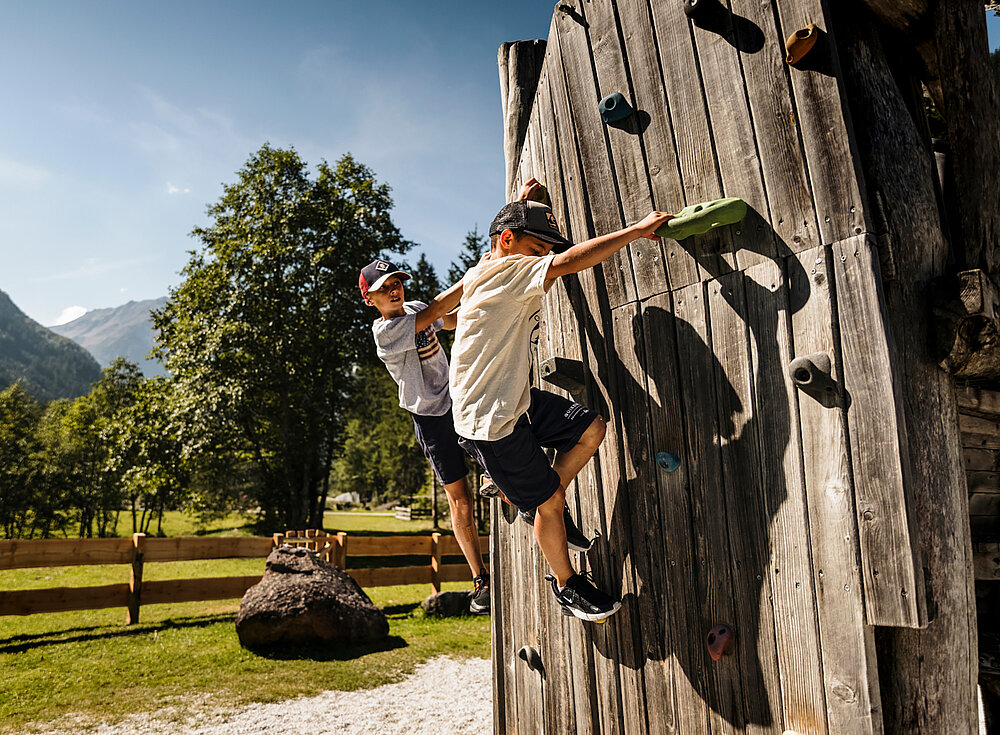 Zwei Jungen klettern an einem sonnigen Tag an einer hölzernen Kletterwand im Freien, im Hintergrund sind Bäume und Berge zu sehen.