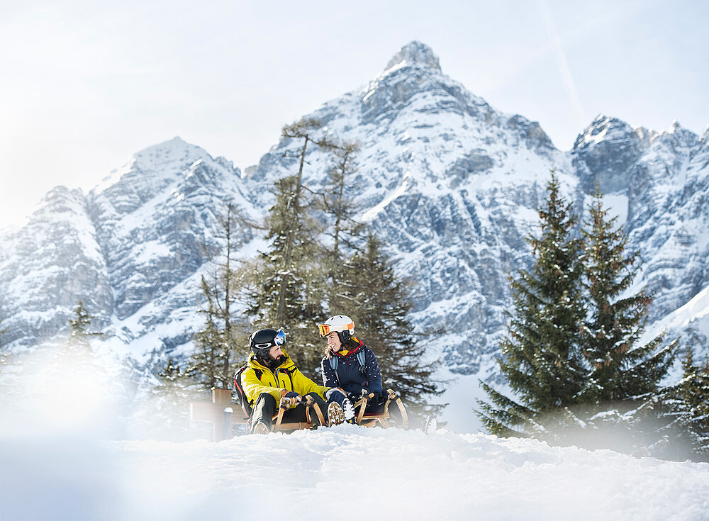Zwei Personen in Winterkleidung sitzen auf Schlitten in schneebedeckten Bergen mit Kiefern und Gipfeln im Hintergrund.